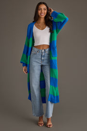 A woman smiles while wearing the Panache Apparel Collection’s Royal Blue & Green Striped Duster Cardigan over a white crop top, paired with light blue high-waisted jeans and brown sandals, standing against a plain grey background.
