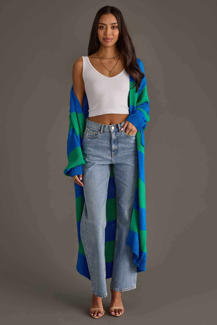 A woman stands against a plain gray background wearing the Panache Apparel Collection Royal Blue & Green Striped Duster Cardigan, paired with a white sleeveless crop top, light blue high-waisted jeans, and sandals.