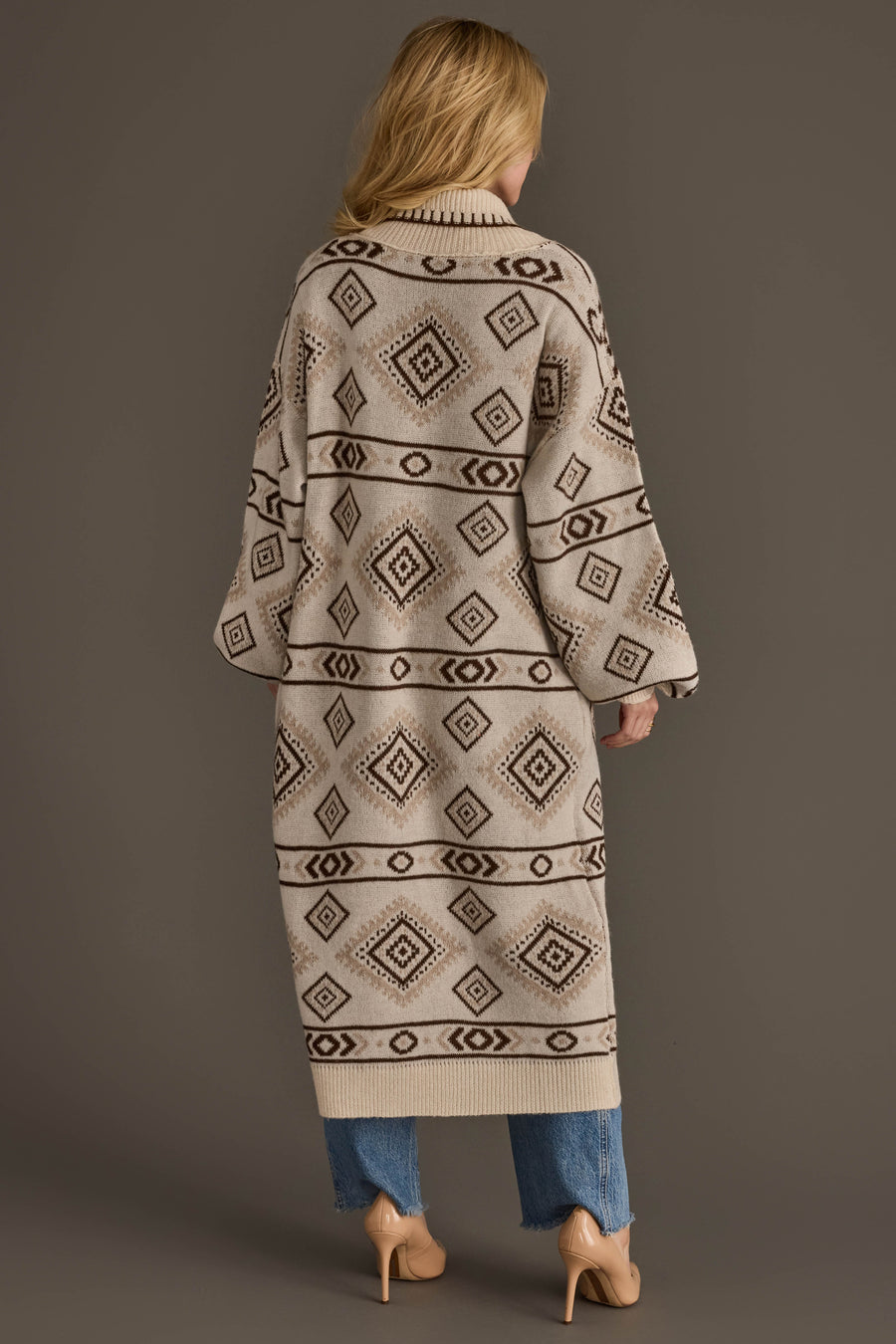 A woman with long blonde hair models the Cream & Brown Western Cardigan from Panache Apparel Collection, pairing it with blue jeans and nude heels. She stands against a plain brown background, facing away from the camera.