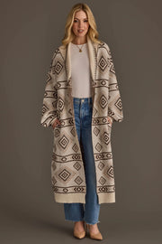 A woman stands against a plain brown background wearing the Panache Apparel Collection Cream & Brown Western Cardigan, a long cardigan featuring a geometric diamond pattern, layered over a white top and blue jeans, paired with tan heels.