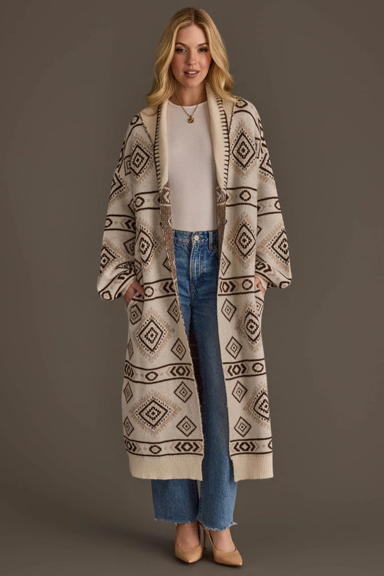 A woman stands against a plain brown background wearing the Panache Apparel Collection Cream & Brown Western Cardigan, a long cardigan featuring a geometric diamond pattern, layered over a white top and blue jeans, paired with tan heels.