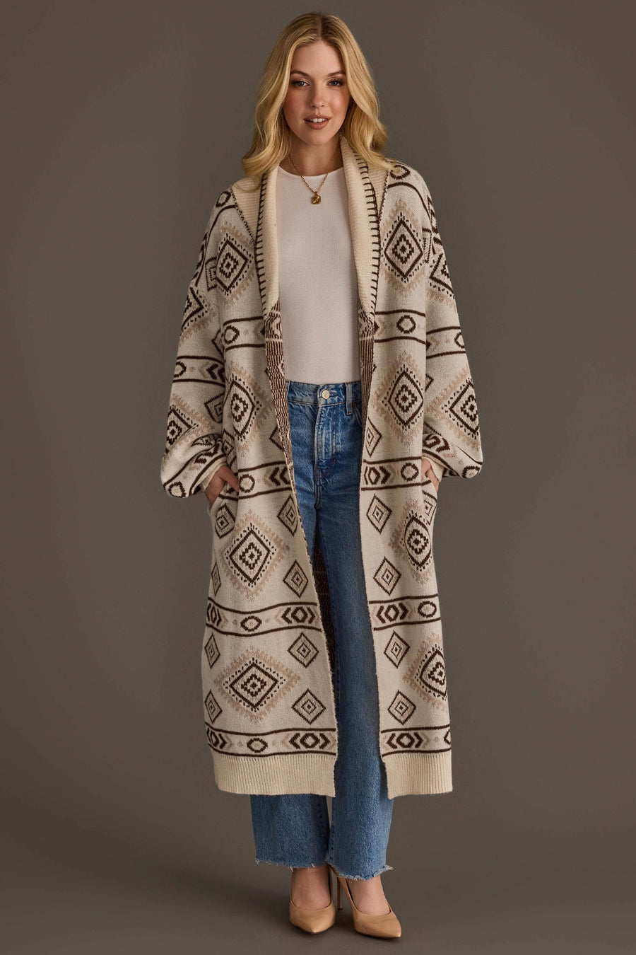 A woman stands against a plain brown background wearing the Panache Apparel Collection Cream & Brown Western Cardigan, a long cardigan featuring a geometric diamond pattern, layered over a white top and blue jeans, paired with tan heels.