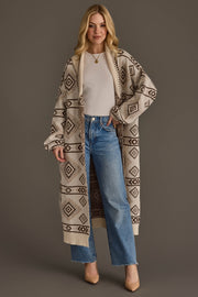 A woman stands against a plain background wearing the Panache Apparel Collection’s Cream & Brown Western Cardigan over a white top, paired with blue high-waisted jeans and tan heels. Her loose blonde hair frames her face as she poses with one hand in her pocket.