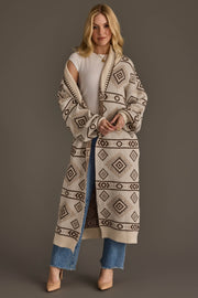 A woman wearing the Panache Apparel Collection’s Cream & Brown Western Cardigan over a white top and blue jeans stands against a plain background, smiling slightly with her hands clasped and beige heels completing her outfit.