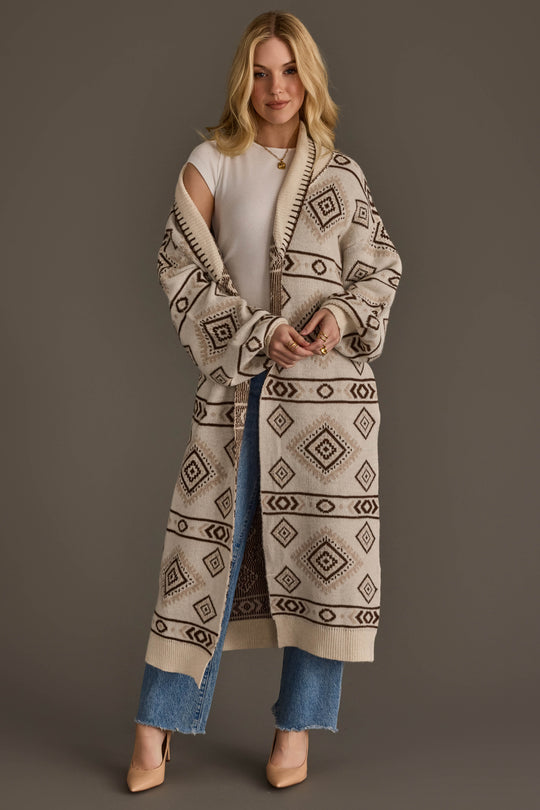 A woman wearing the Panache Apparel Collection’s Cream & Brown Western Cardigan over a white top and blue jeans stands against a plain background, smiling slightly with her hands clasped and beige heels completing her outfit.