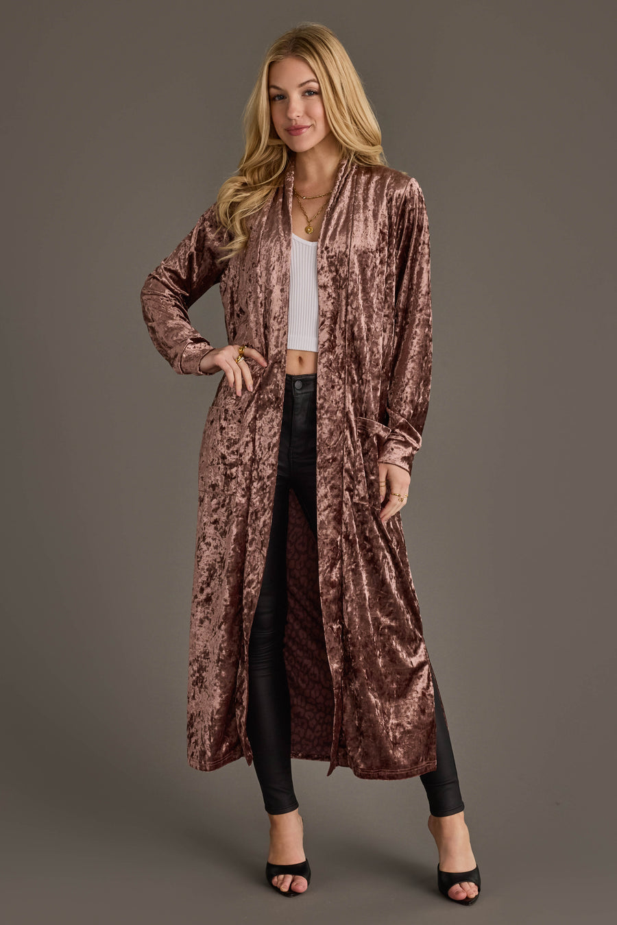 A woman with long blonde hair models the Reddish-Brown Velvet Duster Cardigan by Panache Apparel Collection over a white crop top, black pants, and black open-toe heels against a plain gray background.