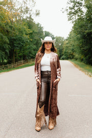 A woman wearing a white cowboy hat, a Reddish-Brown Velvet Duster Cardigan from Panache Apparel Collection, a white top, dark pants, and fringed tan boots stands on an asphalt road.