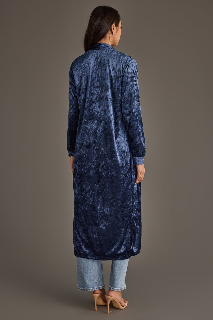 A woman faces away, wearing the Dark Blue Velvet Duster Cardigan by Panache Apparel Collection over light blue jeans and beige heeled sandals, set against a plain neutral background.