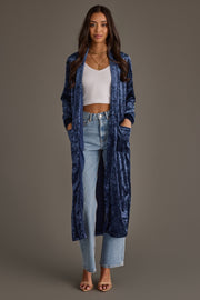 A woman stands facing forward in a Dark Blue Velvet Duster Cardigan by Panache Apparel Collection, paired with a white ribbed crop top, light blue high-waisted jeans, and heeled sandals against a plain brown background.