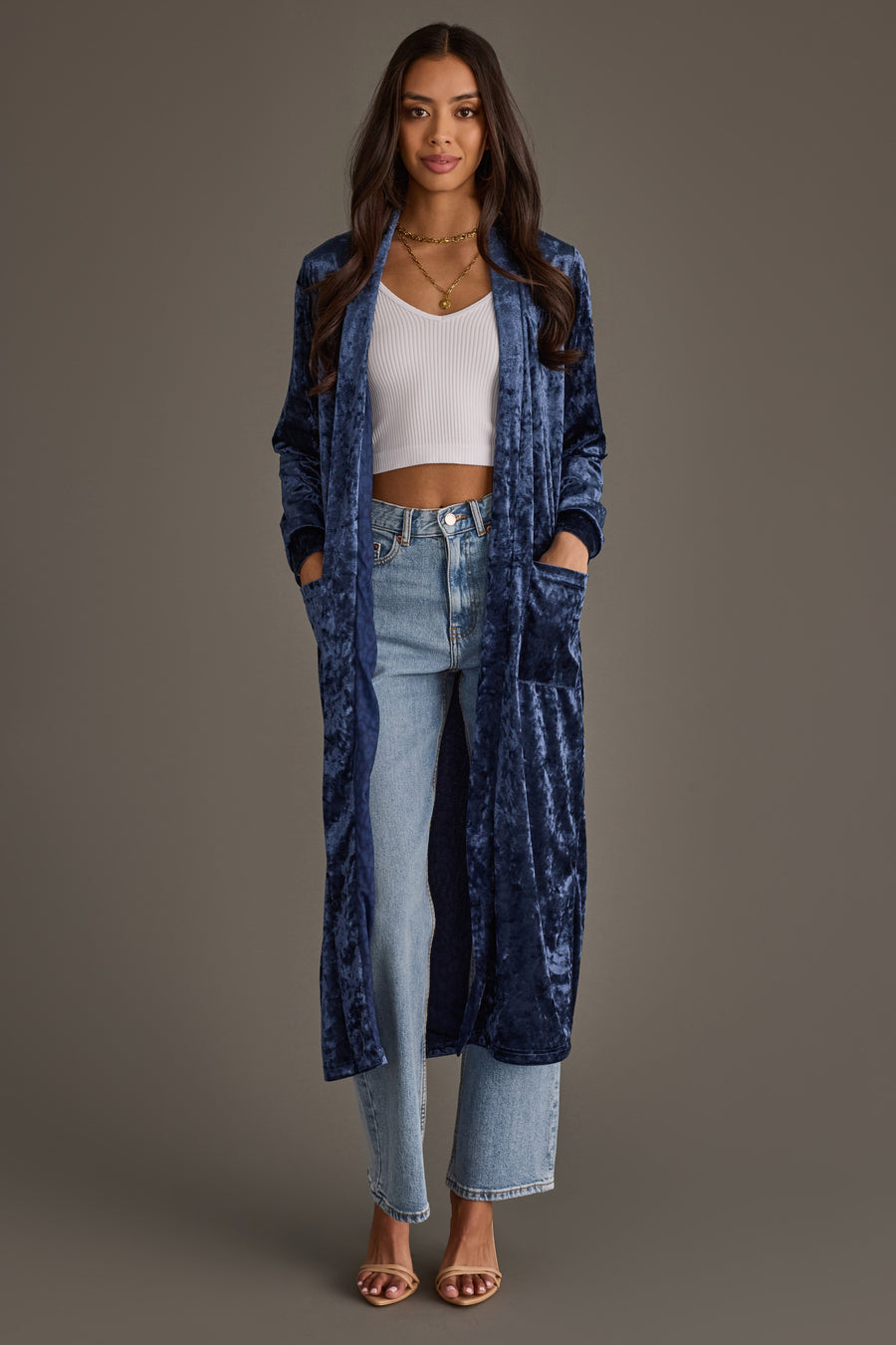 A woman stands facing forward in a Dark Blue Velvet Duster Cardigan by Panache Apparel Collection, paired with a white ribbed crop top, light blue high-waisted jeans, and heeled sandals against a plain brown background.