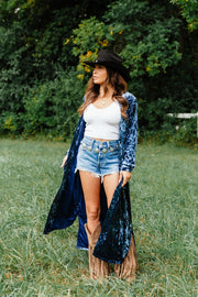 A woman wearing a Dark Blue Velvet Duster Cardigan from Panache Apparel Collection over a white tank top and denim shorts, with a cowboy hat and fringed boots, standing in a grassy field.