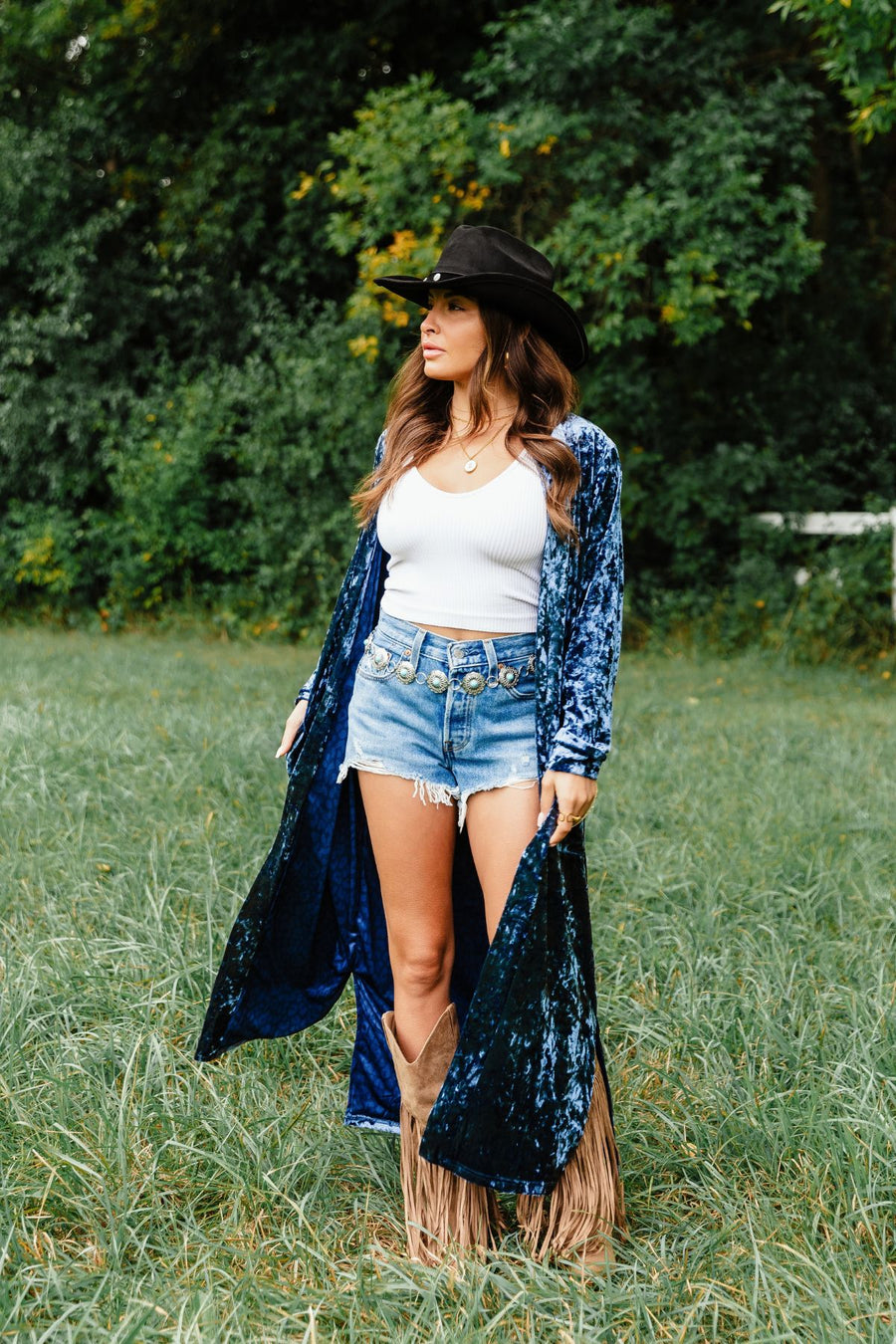 A woman wearing a Dark Blue Velvet Duster Cardigan from Panache Apparel Collection over a white tank top and denim shorts, with a cowboy hat and fringed boots, standing in a grassy field.