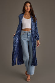 A woman wears the Dark Blue Velvet Duster Cardigan by Panache Apparel Collection over a white crop top, high-waisted light blue jeans, and sandals against a plain gray background.