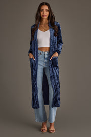 Woman wearing the Dark Blue Velvet Duster Cardigan by Panache Apparel Collection over a white crop top, light blue high-waisted jeans, and heeled sandals; hands in pockets, neutral brown background.