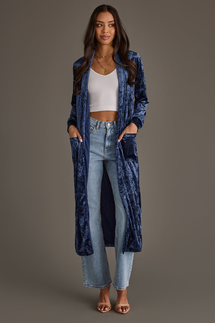 Woman wearing the Dark Blue Velvet Duster Cardigan by Panache Apparel Collection over a white crop top, light blue high-waisted jeans, and heeled sandals; hands in pockets, neutral brown background.