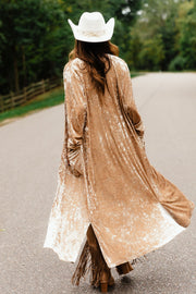 A person wearing a tan Tan Velvet Duster Cardigan, a white cowboy hat, and fringed boots walking down a road.