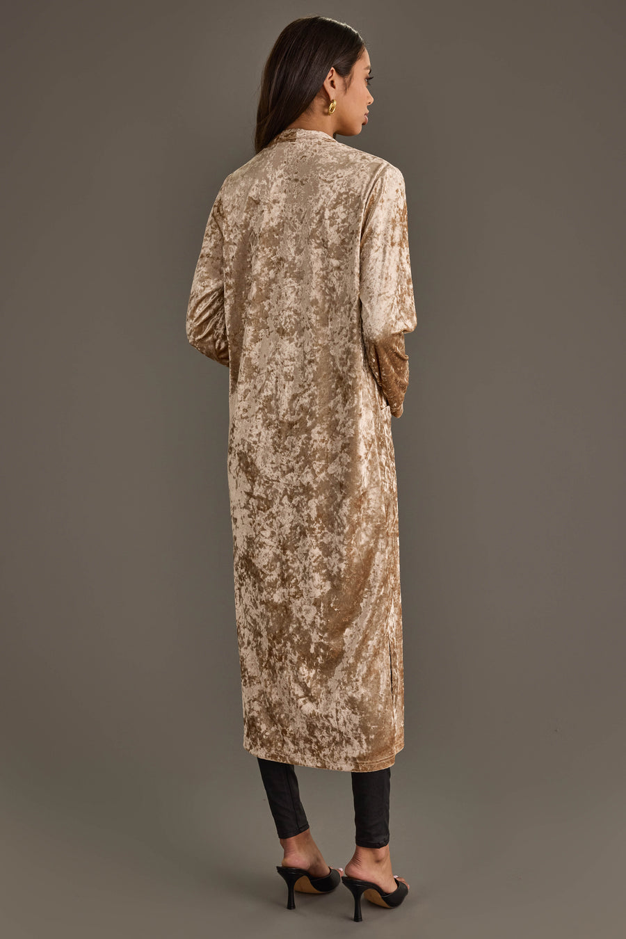 A woman faces away, wearing the Tan Velvet Duster Cardigan by Panache Apparel Collection with black pants and heeled sandals, against a plain grey background.