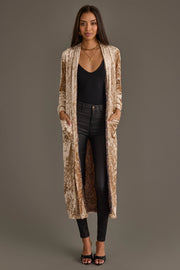 A woman models the Panache Apparel Collection Tan Velvet Duster Cardigan over a black top and skinny jeans, paired with black heels and layered necklaces, hands in the cardigan’s pockets.