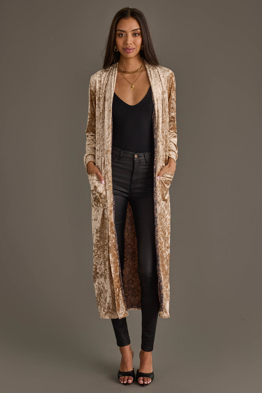 A woman models the Panache Apparel Collection Tan Velvet Duster Cardigan over a black top and skinny jeans, paired with black heels and layered necklaces, hands in the cardigan’s pockets.