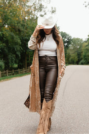 A woman wearing a Tan Velvet Duster Cardigan from the Panache Apparel Collection, white bodysuit, dark pants, a white cowboy hat, and fringed boots walks down a road.