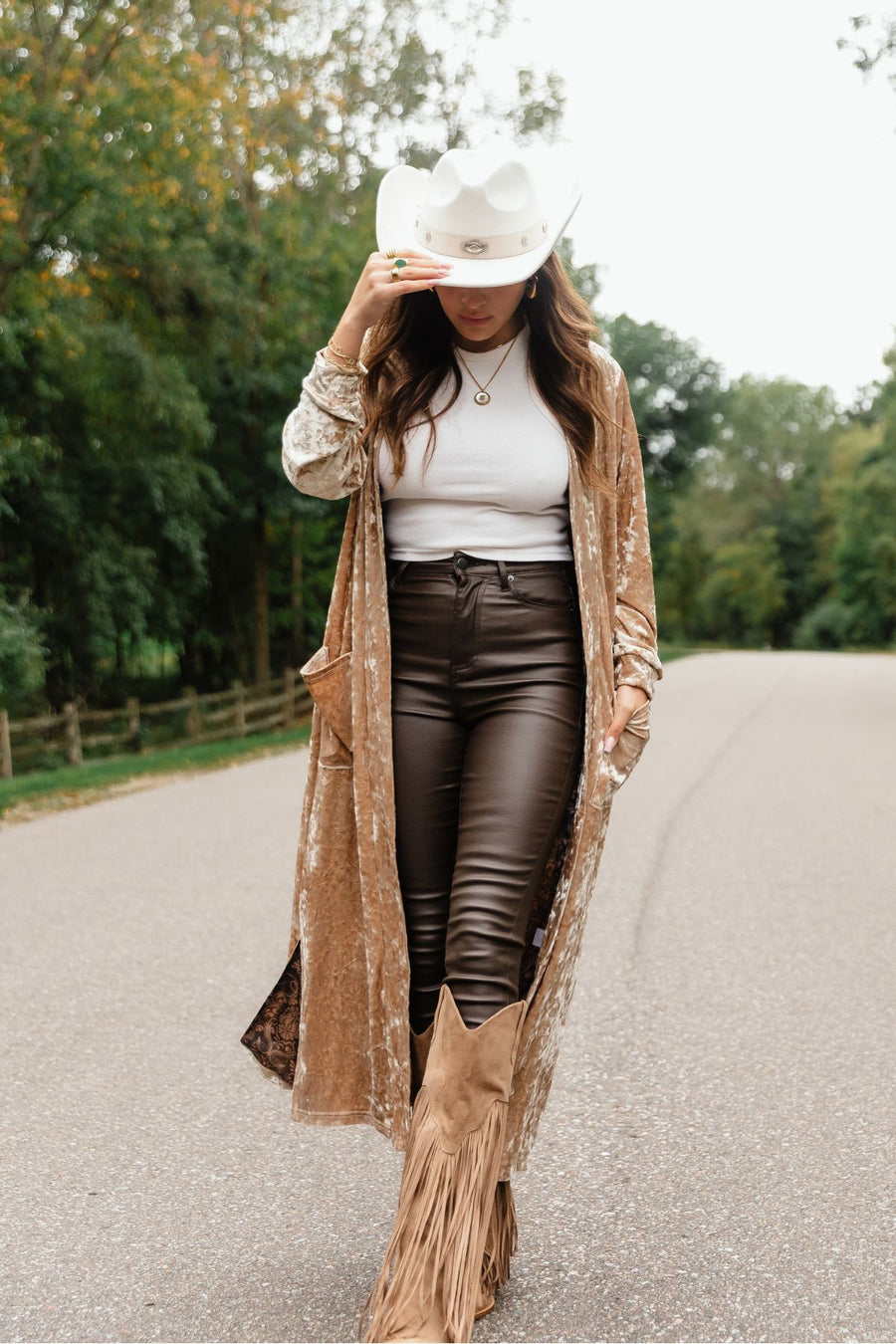 A woman wearing a Tan Velvet Duster Cardigan from the Panache Apparel Collection, white bodysuit, dark pants, a white cowboy hat, and fringed boots walks down a road.