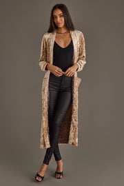 A woman in the Panache Apparel Collection Tan Velvet Duster Cardigan, paired with a black top, black leather pants, open-toe heels, and gold jewelry, stands against a plain background.