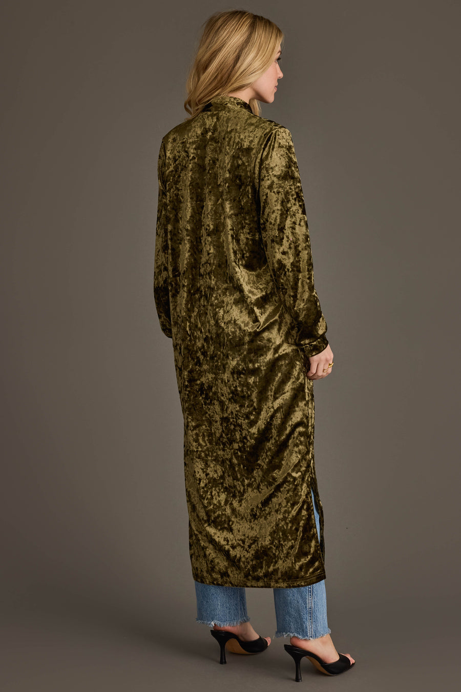 A woman faces away, wearing the Olive Velvet Duster Cardigan by Panache Apparel Collection over blue jeans and black open-toe heels. Its subtle shine, side slit, and her loose blonde hair complete the look.