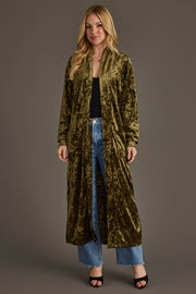 A woman models the Olive Velvet Duster Cardigan by Panache Apparel Collection over a black top, blue jeans, and black open-toe heels against a plain brown background.