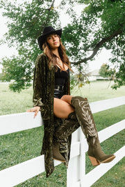 A woman wearing a black hat, an Olive Velvet Duster Cardigan from Panache Apparel Collection, and olive green knee-high boots sits on a white fence.