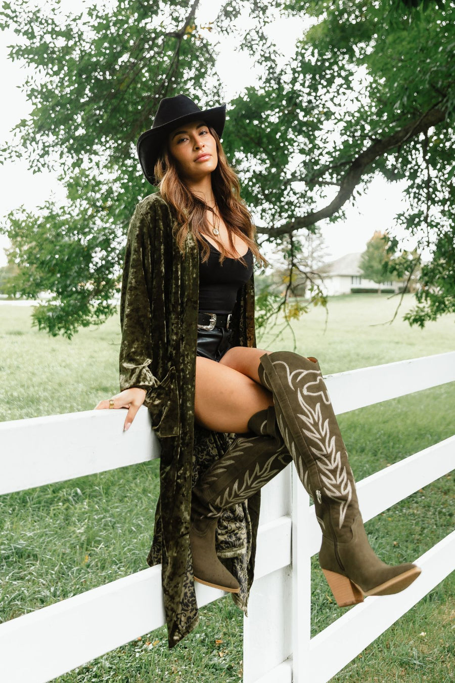 A woman wearing a black hat, an Olive Velvet Duster Cardigan from Panache Apparel Collection, and olive green knee-high boots sits on a white fence.