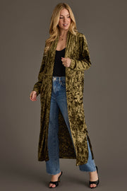 A woman wearing the Olive Velvet Duster Cardigan by Panache Apparel Collection over a black top and frayed blue jeans, paired with black open-toe heels, stands against a plain background, smiling to the side.
