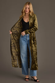 A woman wears the Olive Velvet Duster Cardigan by Panache Apparel Collection over a black top and blue jeans, showing off its patterned lining against a plain background.