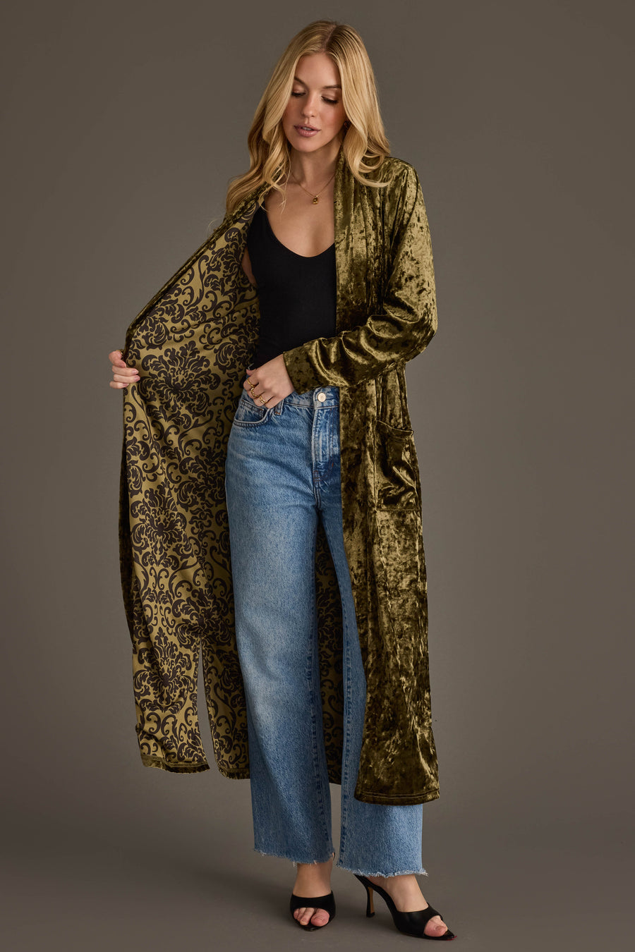 A woman wears the Olive Velvet Duster Cardigan by Panache Apparel Collection over a black top and blue jeans, showing off its patterned lining against a plain background.