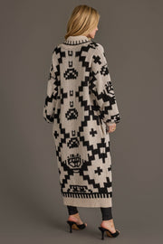 A woman with blonde hair stands facing away, wearing the Panache Apparel Collection’s Cream & Black Western Cardigan, featuring bold black geometric patterns on a beige background. She pairs it with dark pants and heeled sandals for a cozy, stylish look.