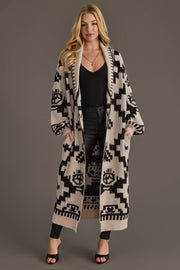 A woman stands facing forward with her hands in the pockets of a Panache Apparel Collection Cream & Black Western Cardigan, worn over a black top and black pants, against a plain gray background.