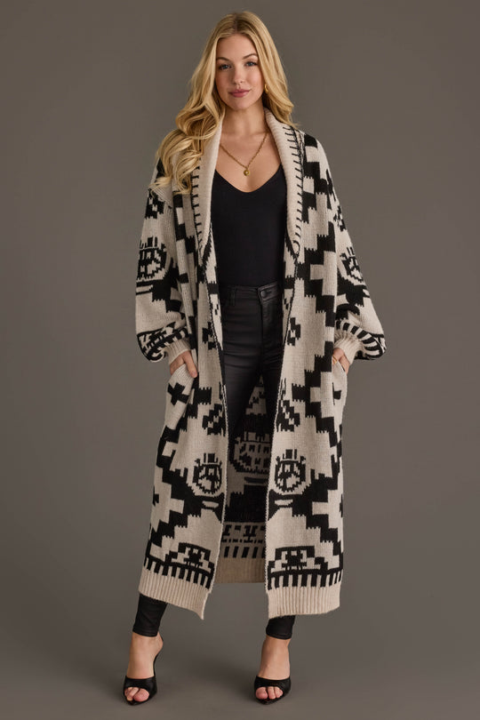 A woman stands facing forward with her hands in the pockets of a Panache Apparel Collection Cream & Black Western Cardigan, worn over a black top and black pants, against a plain gray background.
