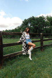A woman with wavy blonde hair wears the Panache Apparel Collection’s Light Blue, Rust, White & Black Western Cardigan featuring geometric patterns, paired with light jeans and beige heeled sandals. She stands against a plain background, facing away from the camera.