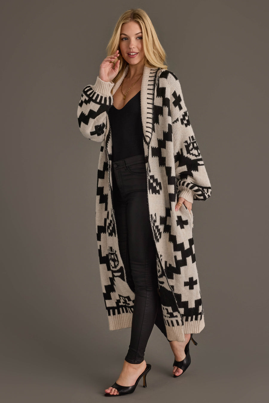 A woman models the Cream & Black Western Cardigan from Panache Apparel Collection, styled over a black top and pants with black heeled sandals, posing against a plain gray background.