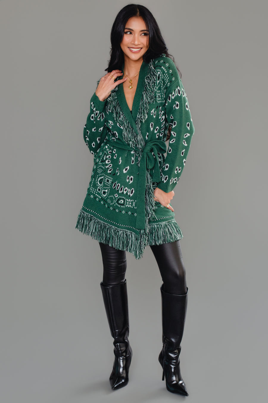 A woman with long dark hair is wearing the Panache Apparel Collection Green Bandana Fringe Cardigan with a belt, paired with black leather pants and gold jewelry, standing against a plain gray background.