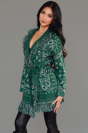 A woman with long dark hair models the Panache Apparel Collection Green Bandana Fringe Cardigan, featuring patterned fabric and fringe at the shoulders and hem, paired with black leggings. She is standing against a plain gray background, facing away from the camera.