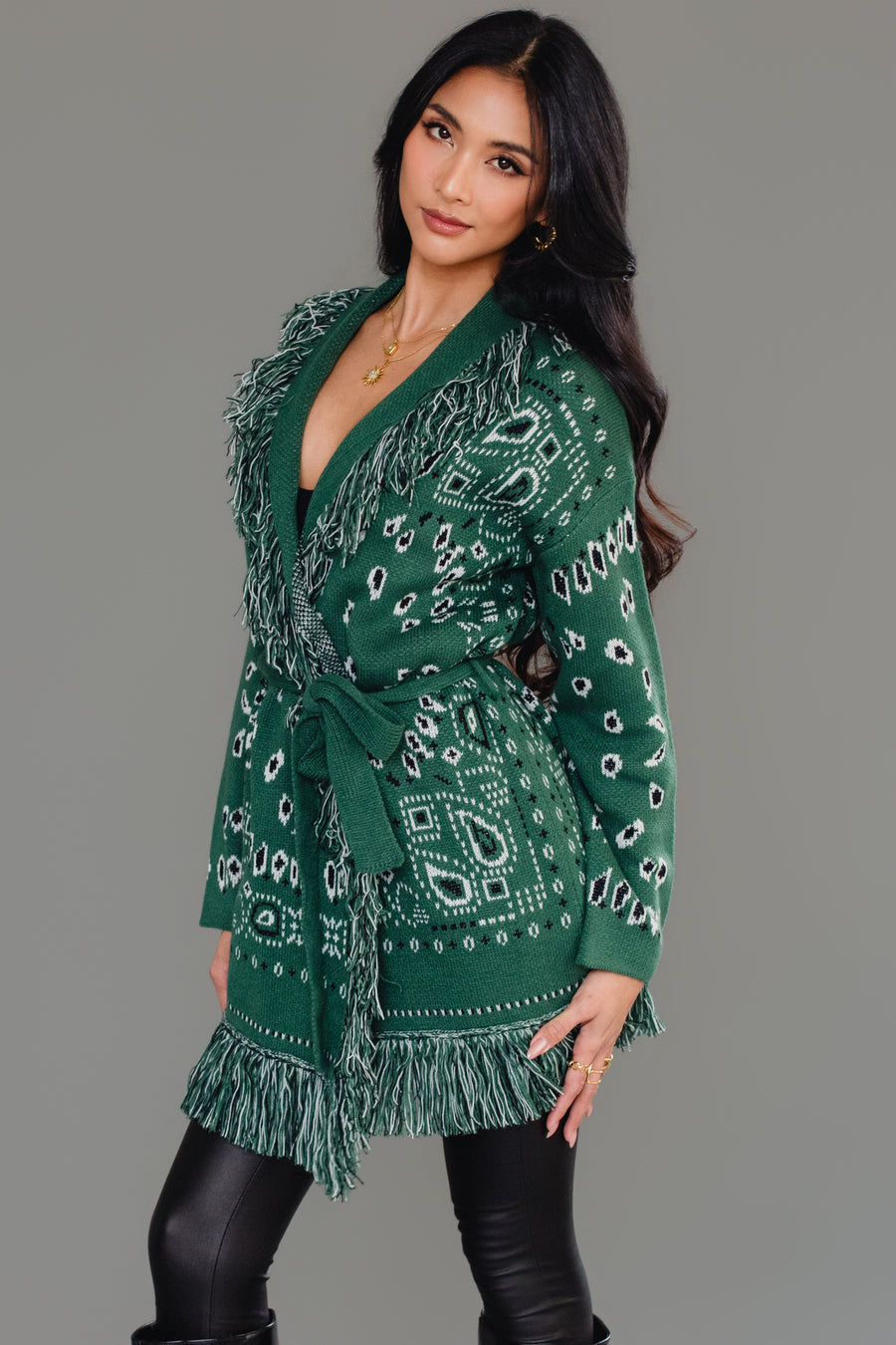 A woman with long dark hair models the Panache Apparel Collection Green Bandana Fringe Cardigan, featuring patterned fabric and fringe at the shoulders and hem, paired with black leggings. She is standing against a plain gray background, facing away from the camera.