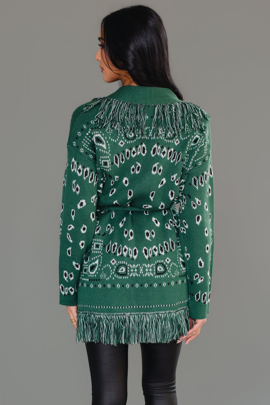 A woman models the Panache Apparel Collection Green Bandana Fringe Cardigan, paired with a black top, black leather pants, and black knee-high boots, standing against a gray background.
