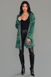 A woman wearing the Panache Apparel Collection’s Blue Bandana Fringe Cardigan, a belted, fringe-trimmed piece featuring a paisley motif, layered over a white top and light blue jeans, stands smiling against a plain background.