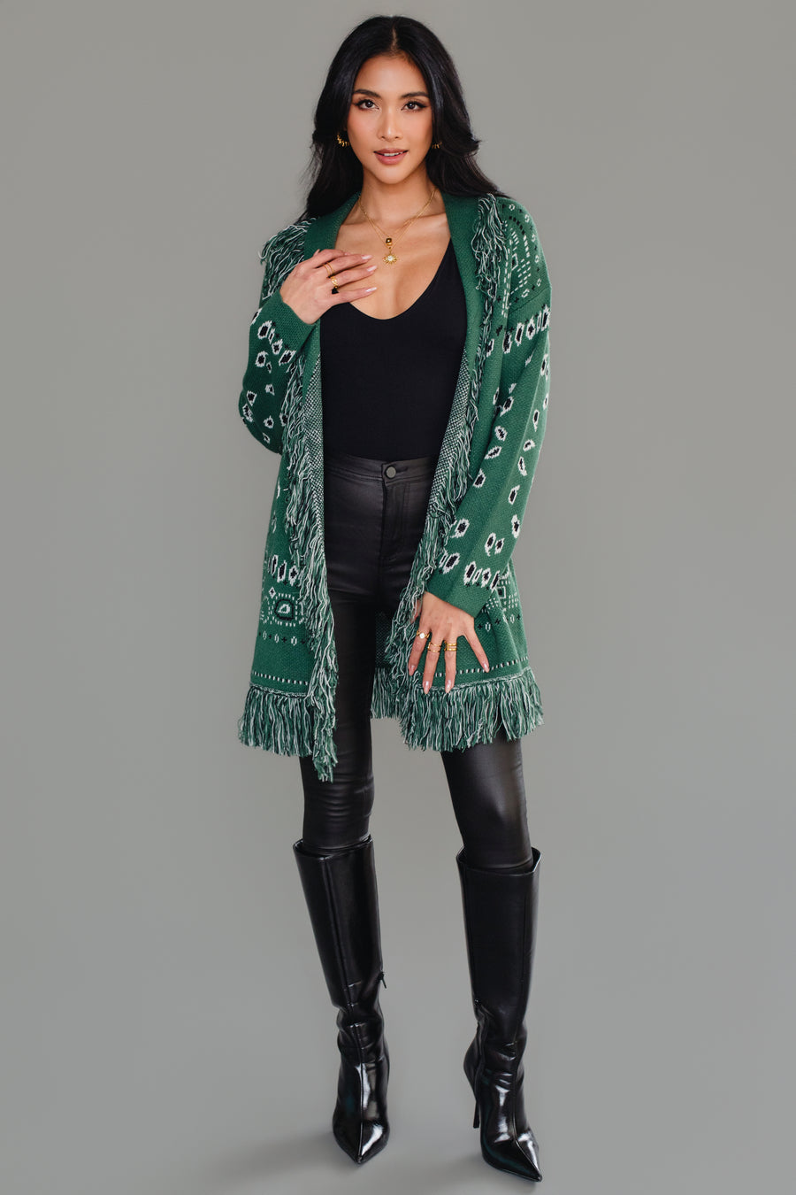 A woman wearing the Panache Apparel Collection’s Blue Bandana Fringe Cardigan, a belted, fringe-trimmed piece featuring a paisley motif, layered over a white top and light blue jeans, stands smiling against a plain background.