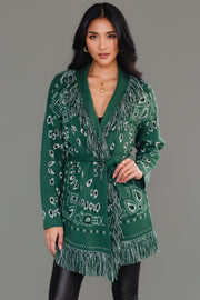 A woman poses against a plain gray background wearing the Panache Apparel Collection Green Bandana Fringe Cardigan, styled with its matching belt, black shiny leggings, and knee-high black boots.