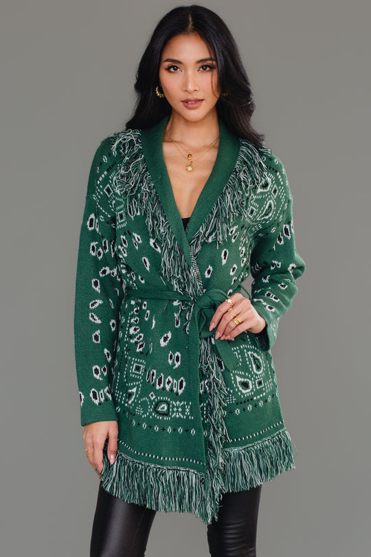A woman poses against a plain gray background wearing the Panache Apparel Collection Green Bandana Fringe Cardigan, styled with its matching belt, black shiny leggings, and knee-high black boots.
