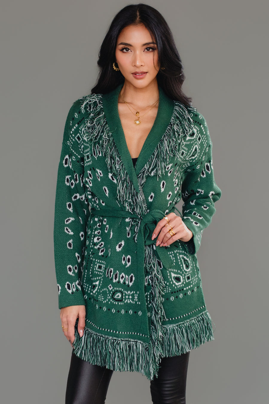 A woman poses against a plain gray background wearing the Panache Apparel Collection Green Bandana Fringe Cardigan, styled with its matching belt, black shiny leggings, and knee-high black boots.