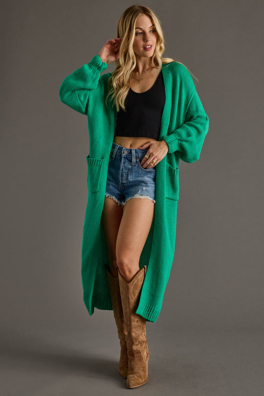 A woman models the Hilton Duster Cardigan in green by Panache Apparel Collection, styled with a black crop top, denim shorts, and tan cowboy boots against a plain gray background for a cozy layered look.