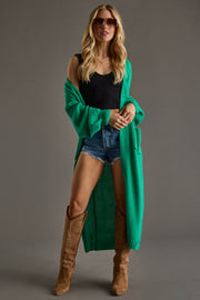 A woman wearing sunglasses, the Panache Apparel Collection Hilton Duster Cardigan in green over a black tank top, denim shorts, and tan knee-high boots stands against a neutral background.