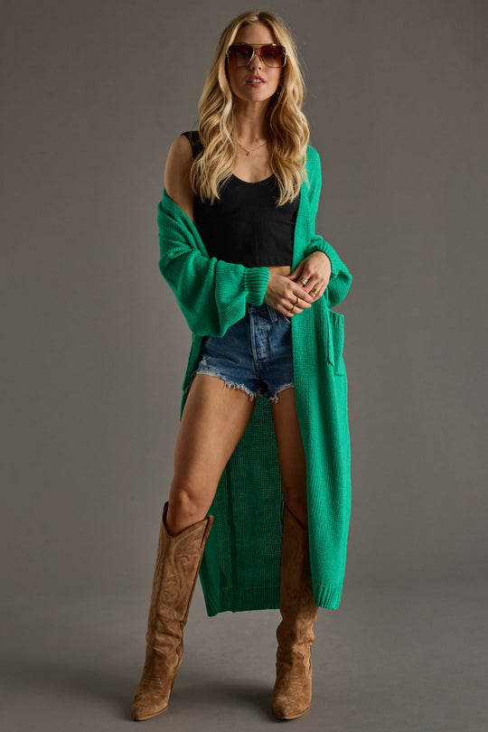 A woman wearing sunglasses, the Panache Apparel Collection Hilton Duster Cardigan in green over a black tank top, denim shorts, and tan knee-high boots stands against a neutral background.
